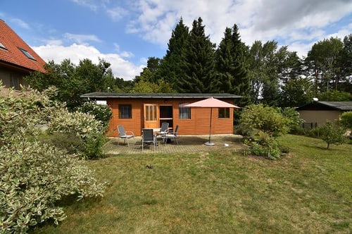 Braunes Holzhaus mit Terrasse, Gartenmöbeln und rotem Sonnenschirm auf einer grünen Wiese.