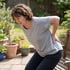 A woman in a garden winces while holding her lower back and standing up from a wooden bench.