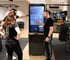 Ein Trainer erklärt zwei Personen Trainingsdaten auf einem großen Milon-Touchscreen in einem modernen Fitnessstudio.