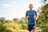 A senior man in a blue t-shirt and headphones jogs outdoors on a sunny park trail.