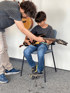 An adult male teaches a young boy to play an electric guitar. The boy sits on a chair with his foot on a small stool.