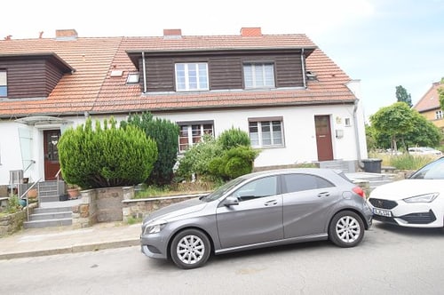 Zwei Autos parken vor einer Reihe von Reihenhäusern mit orangefarbenen Dächern und grünen Büschen.