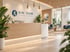 Moderner Empfangsbereich der Kinesium Physiotherapie mit Holztheke, Logo an der Wand und hellem Wartezimmer mit Pflanzen.
