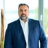 Antonio Benavides, seriöses Business-Porträt eines Mannes im dunklen Anzug mit offenem Hemd vor einem modernen Hintergrund.