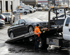 A tow truck operator loads a dark sedan onto a hydraulic tilt-bed tow truck in a parking lot.