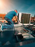 Techniker kniet auf einem Dach und arbeitet an einer Klimaanlage, umgeben von Kabeln und Werkzeugen.