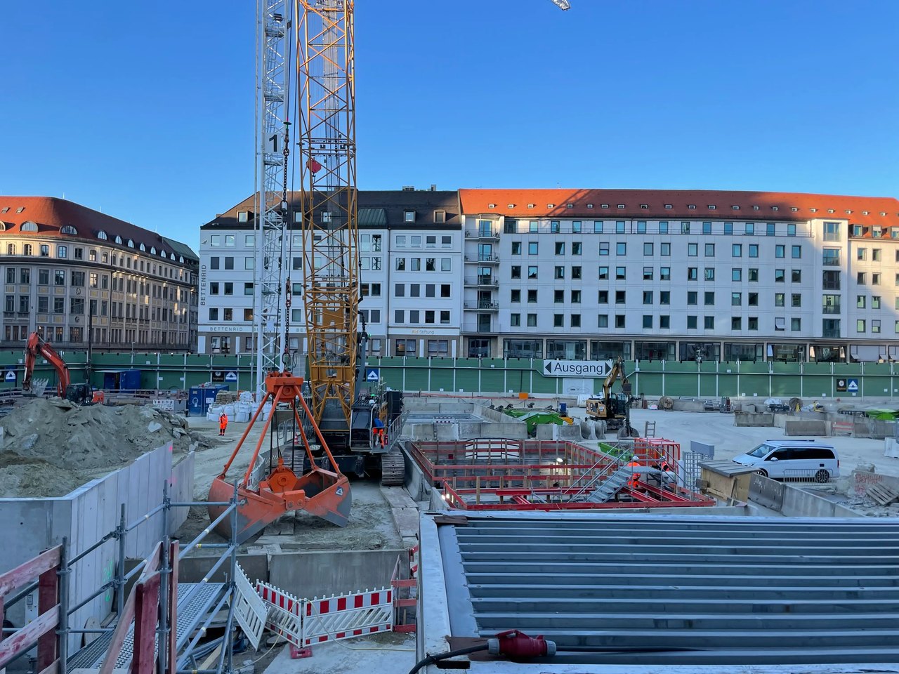 Baustelle mit großem Kran, Erdaushub, Betonwänden und Gebäuden im Hintergrund unter blauem Himmel.