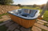 Koller Whirl Systems Hot-Tub: handgefertigter ovaler Holz-Outdoor-Whirlpool mit Edelstahlofen, sprudelndem Wasser und Gartenlounge beim Sonnenuntergang.
