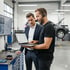 Zwei Männer betrachten einen Laptop in einer Autowerkstatt, ein Auto ist auf einer Hebebühne im Hintergrund.