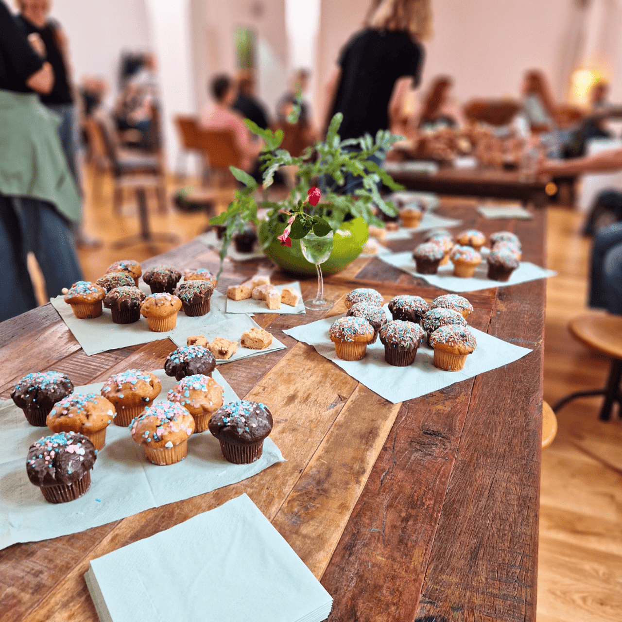 Braune Holztischplatte mit vielen Muffins und Cookies, einer Blumendekoration und verschwommenen Menschen im Hintergrund.