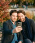 Zwei Männer machen ein Selfie auf einer Parkbank, im Hintergrund Bäume und verschwommene Personen.