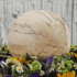 Helle, gemaserte Holzkugel umgeben von einem Kranz aus bunten Blumen und Grünpflanzen vor einer Steinmauer.