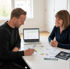 Zwei Personen sitzen an einem Tisch und besprechen Dokumente und einen Laptop, der eine Tabelle anzeigt.
