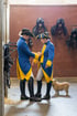 Zwei Frauen in blau-gelben Uniformen mit Dreispitzhüten stehen neben einem Hund in einem Pferdestall mit Sätteln an der Wand.