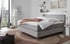 Graues Boxspringbett mit Federkernmatratze, weißer Bettwäsche, brauner Decke und Kissen in einem Schlafzimmer.