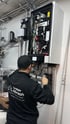 Techniker in schwarzer Arbeitskleidung installiert ein Heizungssystem mit vielen Rohren und Kabeln.