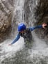 Person in Neoprenanzug und blauem Helm unter einem Wasserfall beim Canyoning.