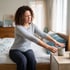 A woman with curly hair sits on a bed and winces while reaching for a mug on a nightstand.