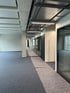 Leerer Büroraum mit grauem Teppichboden, Säulen, glänzenden Deckenleuchten und teilweise abgedecktem Gehweg.