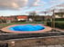 Ein runder Pool mit blauer, aufgewölbter Poolabdeckung und Holzumrandung in einem Garten vor Wohngebäuden.