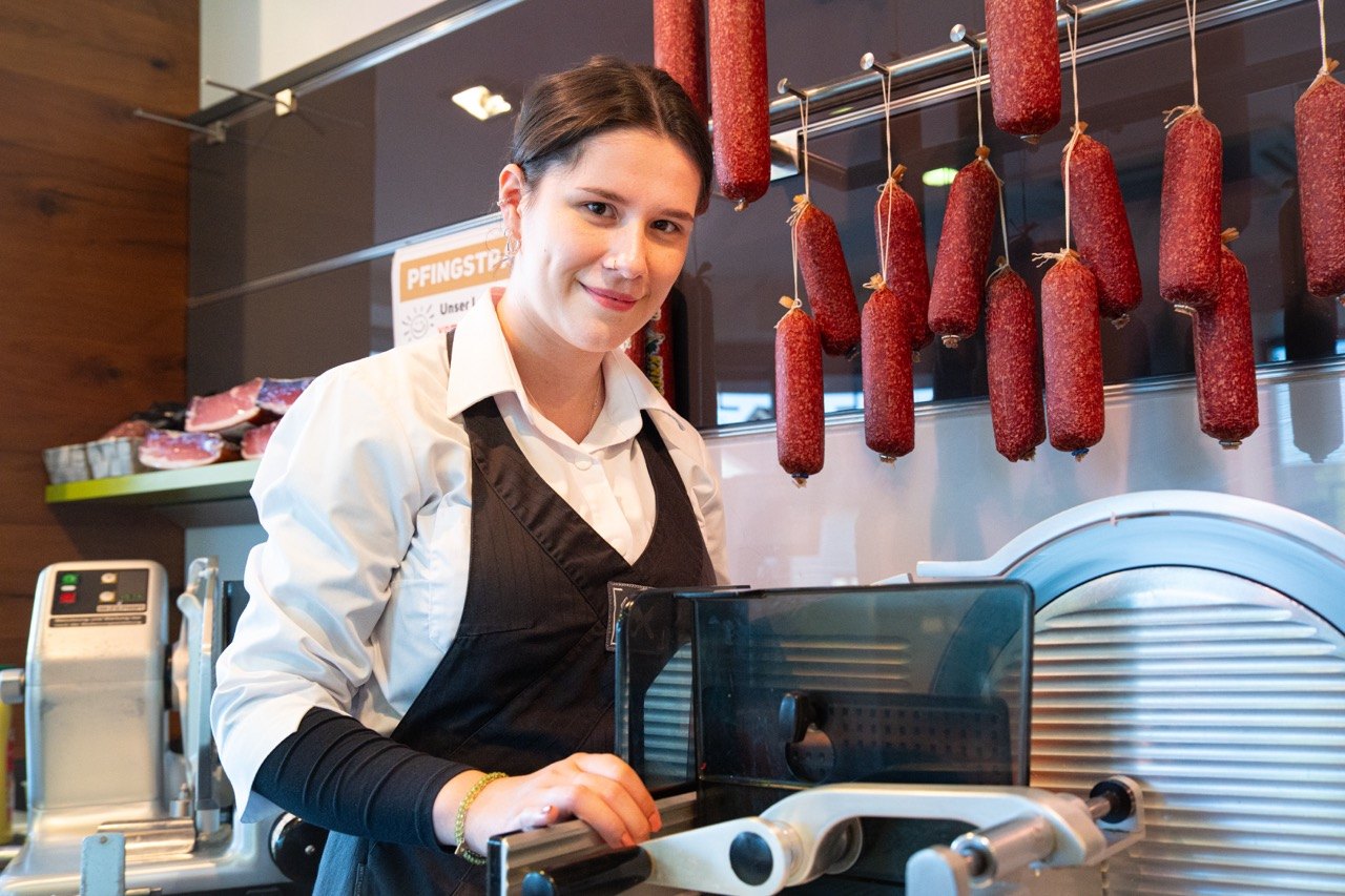 Verkäuferin an der Aufschnittmaschine, dahinter hängende Salami