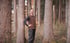 Ein lächelnder Mann mit brauner Weste steht im Wald zwischen hohen Bäumen.