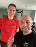 Zwei lächelnde Männer im Fitnessstudio, einer im roten Shirt, der andere im schwarzen Shirt mit der Aufschrift „MAX Personal Trainer“.