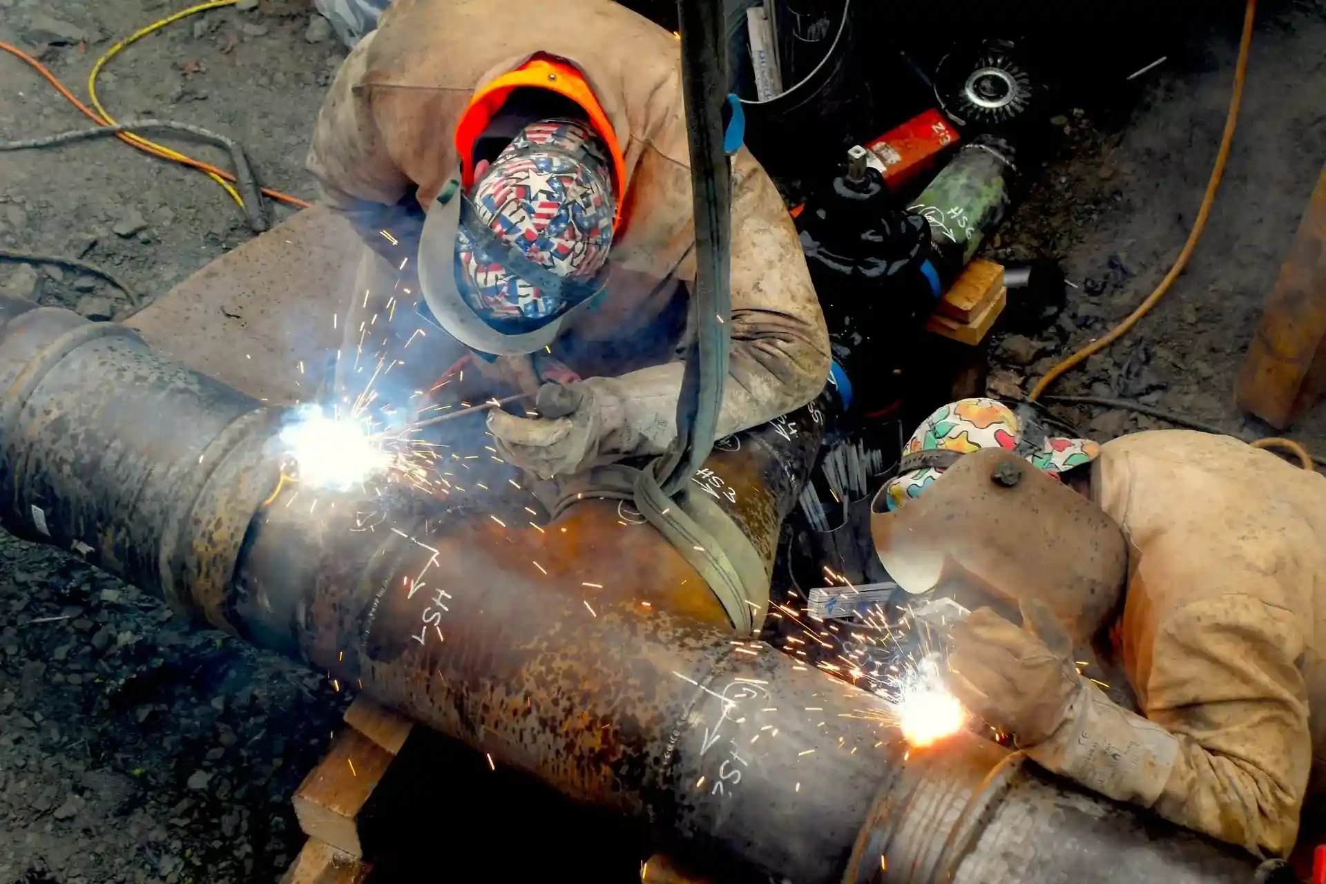Zwei Schweißer der ASK verbinden ein großes Metallrohr, Funken sprühen beim Schweißen.