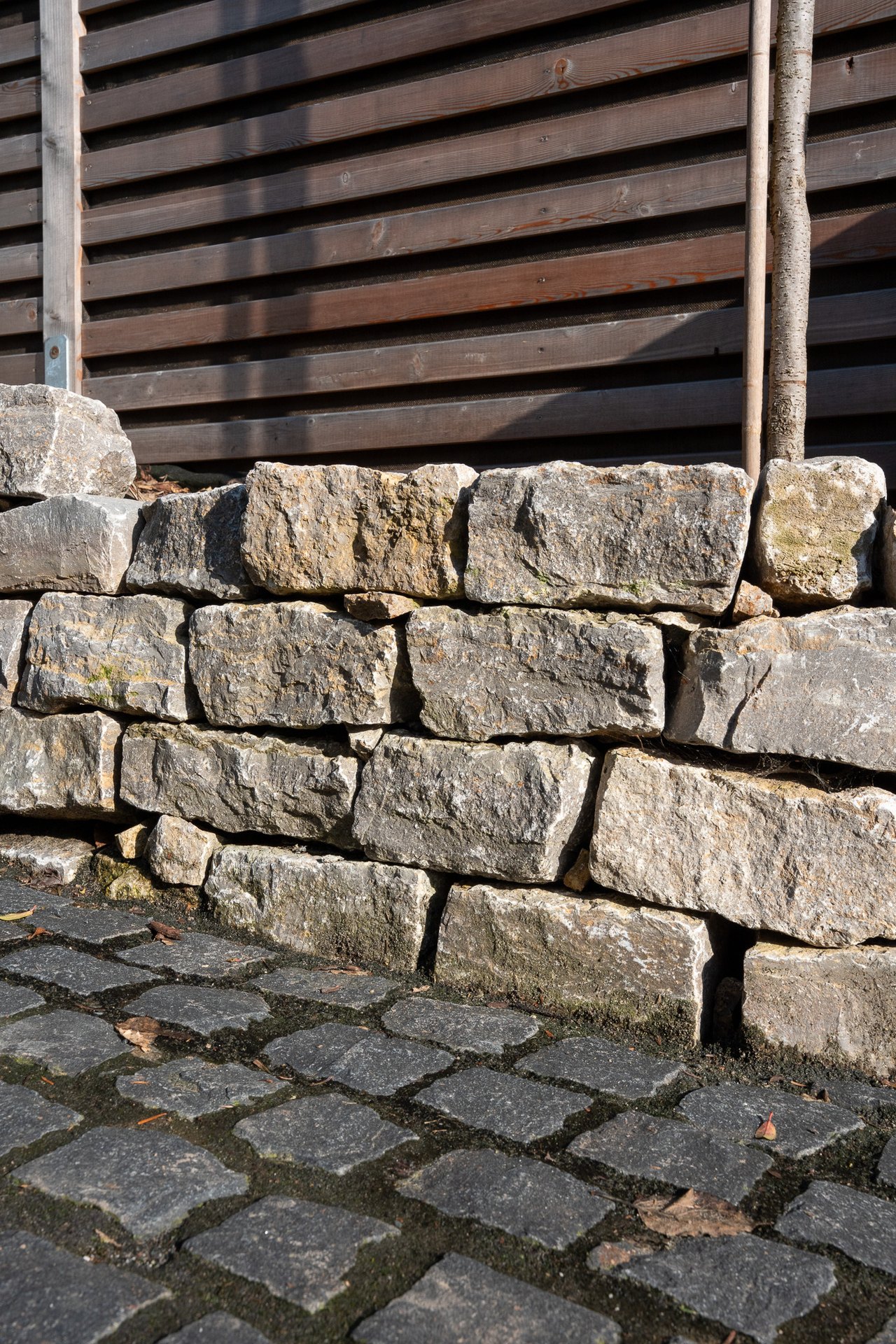 Trockenmauer aus geschichteten Natursteinen mit dunklem Holzzaun und Kopfsteinpflaster davor