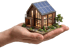 Hand hält ein Miniaturhaus aus Holz mit Solarpanelen auf dem Dach und beleuchteten Fenstern.