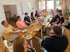 The AlleFarben team sits together at a long breakfast table in a bright room, chatting.