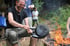 Ein lachender Mann wendet am Lagerfeuer Pfannkuchen in zwei gusseisernen Pfannen.