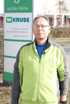 Mann in grün-grauer Jacke und Brille vor einem Schild mit "W KRUSE Ihr Gebäudedienstleister www.wkruse.de".
