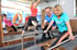 Zwei Frauen trainieren an Fitnessgeräten, während ein Mann einer Frau hilft.
