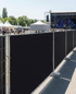 Schwarzer Bauzaun mit Festivalgelände, Bühne und Menschen im Hintergrund.