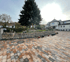 Gepflasterter Vorplatz mit Natursteinmauer, hohem Nadelbaum und einem großen Haus im Hintergrund.
