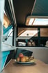 Innenraum eines modern gestalteten Wohnwagens mit Holzelementen, einem Dachfenster und einem Teller mit frischen Früchten wie Äpfeln und Bananen auf dem Tisch.