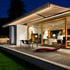 Beige-braun gestreifte Markise mit LED-Beleuchtung über einer Terrasse mit Lounge-Sessel und Glasfassade bei Nacht.