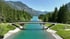 Eine Fußgängerbrücke überspannt türkisfarbenes Wasser zwischen grünen Wäldern und Bergen unter blauem Himmel.