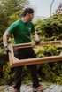 Man in green shirt, black pants, cap, and gloves carrying a wooden frame outdoors.