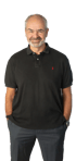 Ein lächelnder Mann mit grauem Bart und Haaren trägt ein schwarzes Poloshirt und eine graue Hose mit Händen in den Taschen.