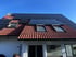 Ziegeldach eines Hauses mit zahlreichen schwarzen Solarmodulen und drei Dachfenstern unter blauem Himmel.