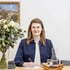 Frau mit Brille und blauem Blazer schreibt an einem Schreibtisch neben einem Blumenstrauß und einer Wasserkaraffe.