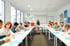 Eine Lehrerin steht vor einer Gruppe von Schülern in einem hellen Klassenzimmer mit Whiteboard und Weltkarte.