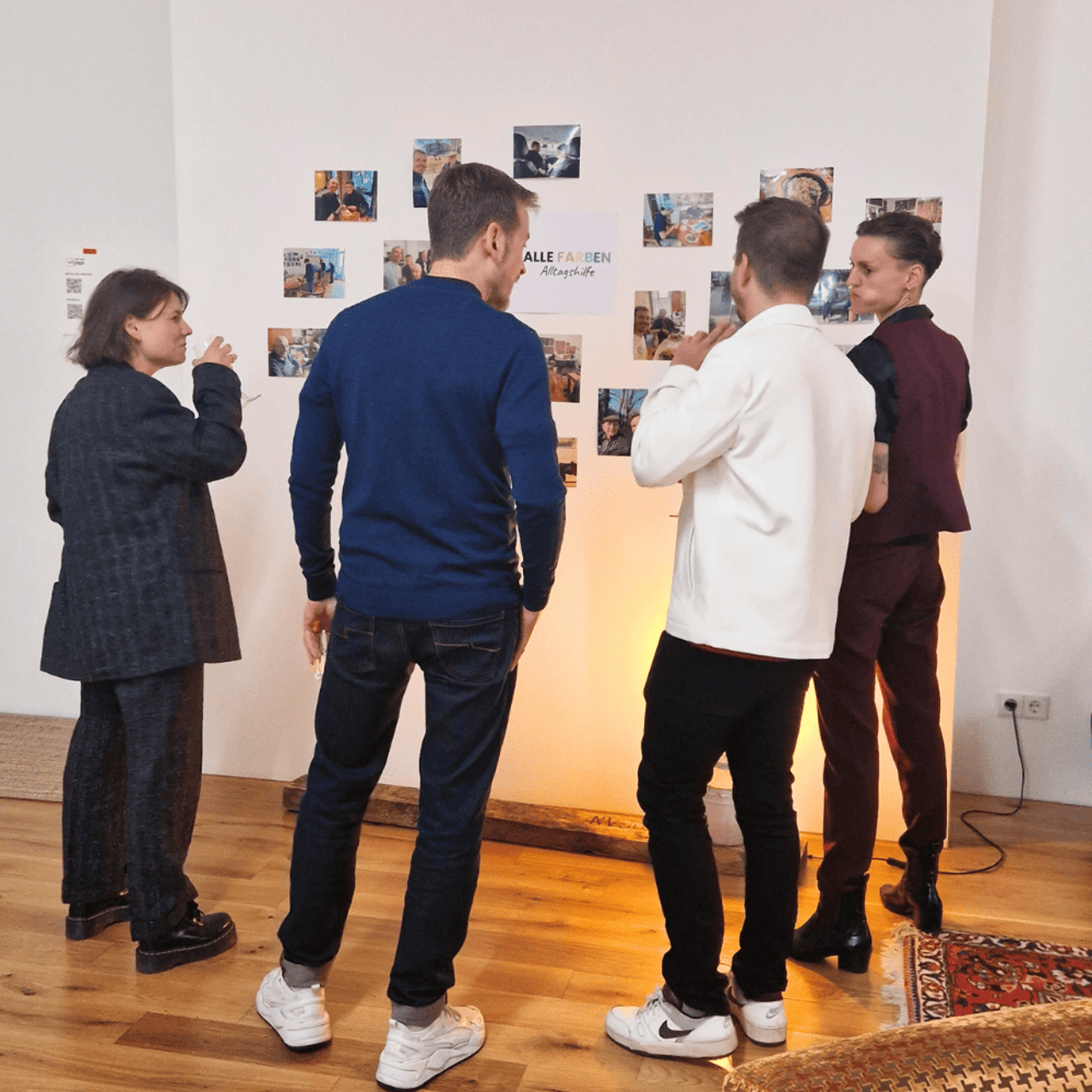 Four people stand in front of a wall with photos and a poster reading “ALLE FARBEN”.
