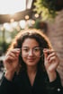 Junge Frau mit lockigem Haar hält eine silberne Brille vor ihr Gesicht, lächelt leicht.