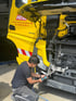 Ein Mechaniker kniet vor einem geöffneten gelben Hess-LKW und arbeitet am Motor.