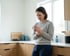A woman in a kitchen holds a mug while gripping her wrist with a pained expression.