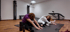 Zwei Frauen führen Dehnübungen auf Matten in einem Fitnessstudio mit Trainingsgeräten und Parkettboden aus.