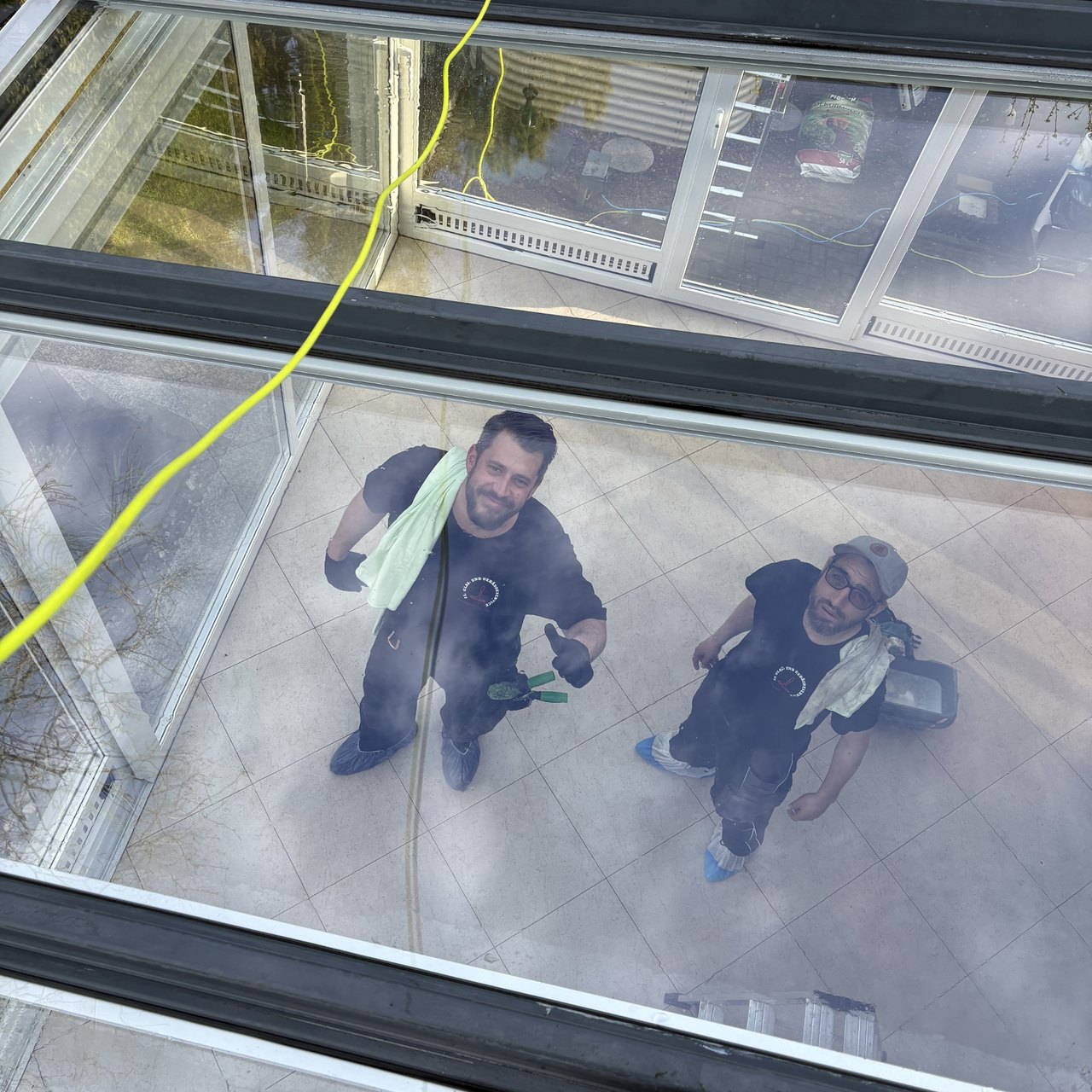 Zwei Fensterputzer in dunkler Arbeitskleidung mit Tüchern blicken durch ein sauberes Glasdach nach oben.
